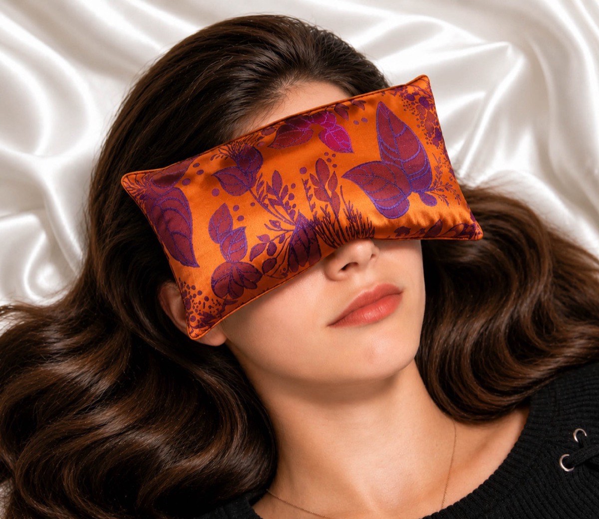 A woman resting with a silk botanical lavender eye pillow across her eyes on white satin sheets